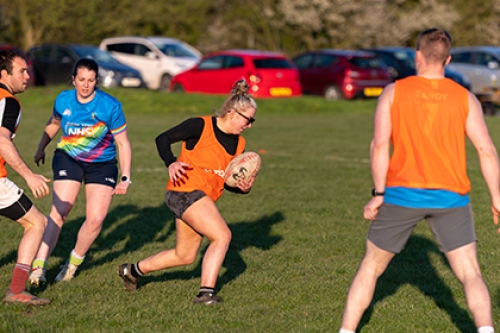 A woman runs as players from both teams look at her during an outdoors rugby game.