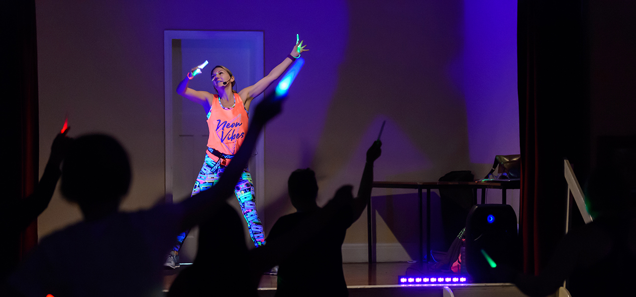 An instructor stands on a stage holding glow sticks as participants follow along with aerobics in front of her