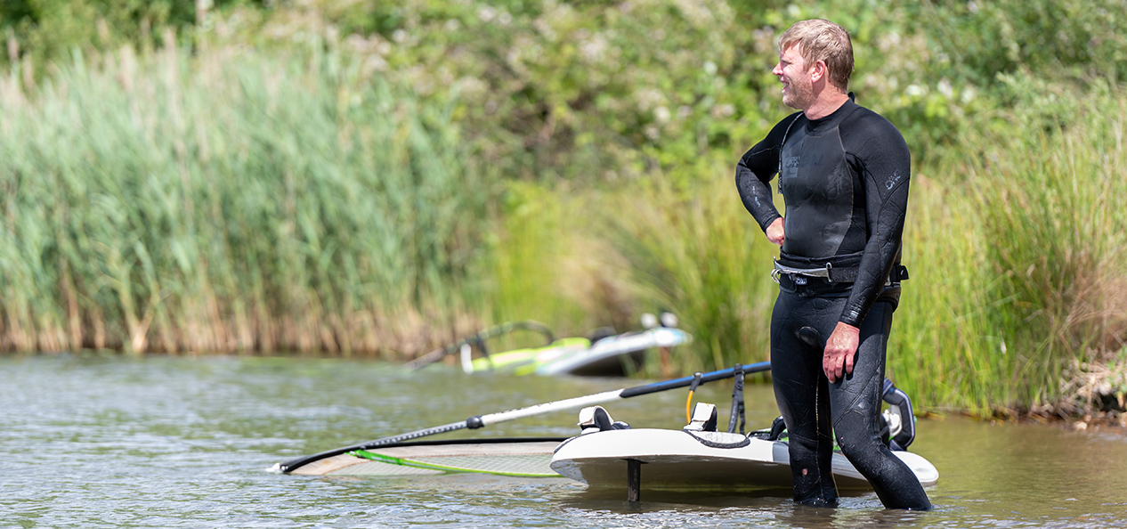 A man in a wetsuit stands in shin-deep water in a lake, next to a windsurfing board and sail