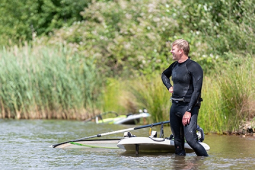 A man in a wetsuit stands in shin-deep water in a lake, next to a windsurfing board and sail