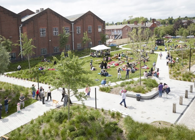 People sitting on rugs on a green space with trees in front of buildings, with others walking on adjacent paths