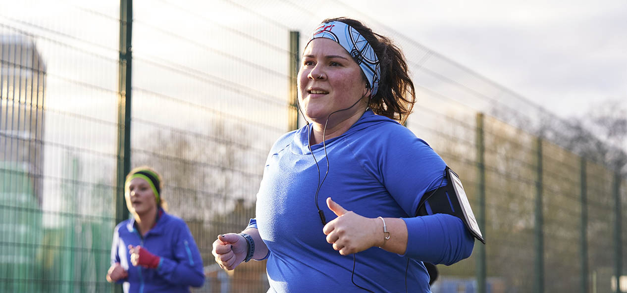 A woman, wearing headphones, runs in a park, with another runner in the background