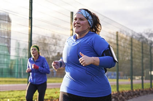 A woman, wearing headphones, runs in a park, with another runner in the background