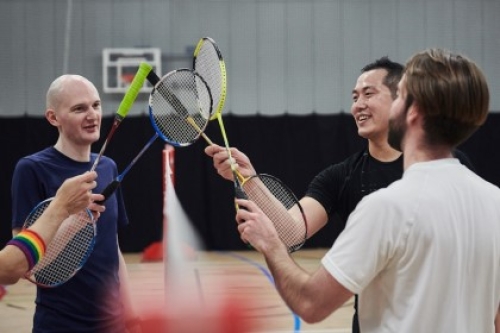 Four people touch their badminton racquets together over the net after a game on an indoor court