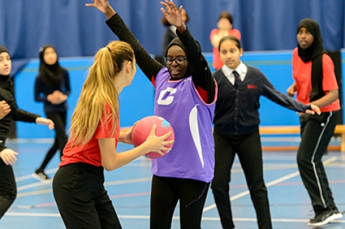 Girls play netball in a PE lesson. One player has the ball and another girl is guarding her, with both arms in the air.