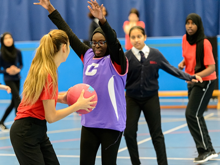 Girls play netball in a school PE lesson. One girl with the ball is guarded by another with both arms in the air.