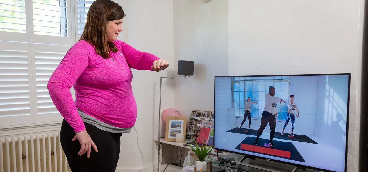 A pregnant woman follows along to an exercise class on her TV