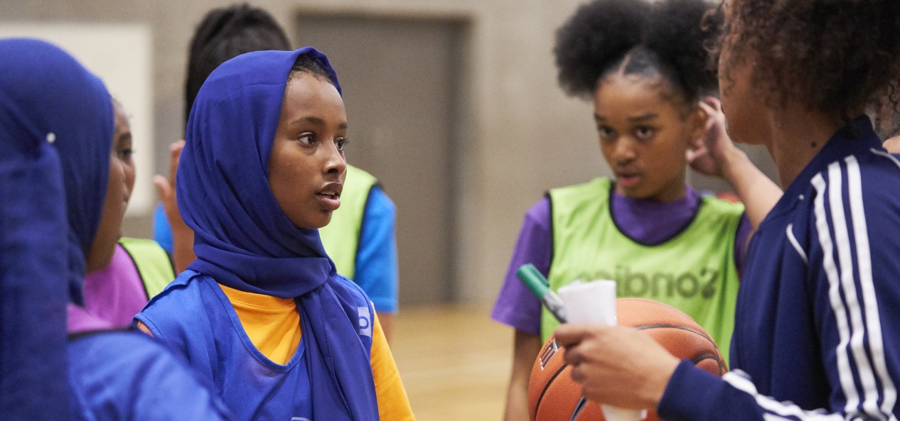 A female coach holding a basketball, pen and paper talks to a group of girls on a court.