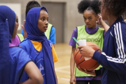 A female coach holding a basketball, pen and paper talks to a group of girls on a court.