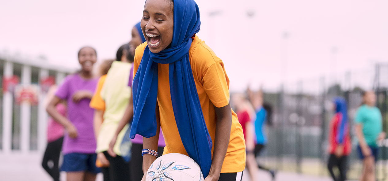 A girl wearing a head scarf laughs and smiles while playing netball in a school playground