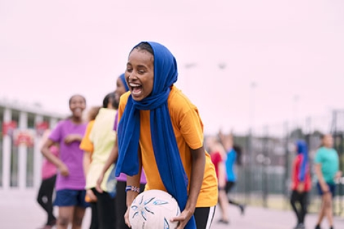 A girl wearing a head scarf laughs and smiles while playing netball in a school playground