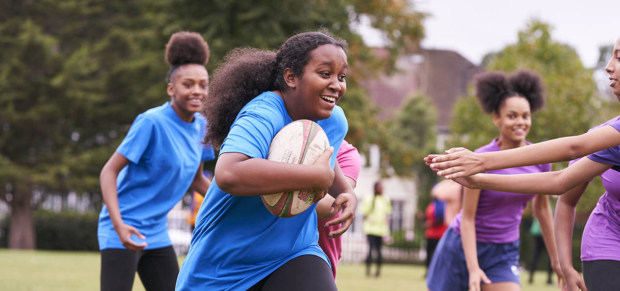 A girl runs with a rugby ball as a group of girls play rugby in a park.