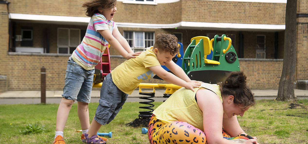 A woman sits on the floor, stretching, while her two children help her by pushing on her back.