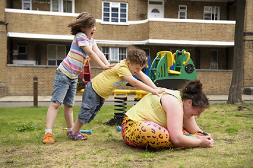 A woman sits on the floor, stretching, while her two children help her by pushing on her back.