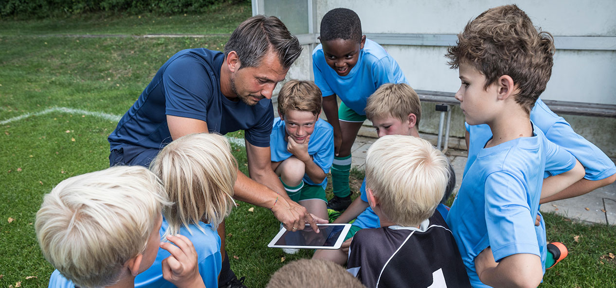 A group of kids listen to the instructions of their coach, who's showing them something on their tablet