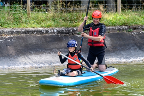 A boy with a prosthetic leg stands on a paddle board, on water. while an instructor kneels behind him