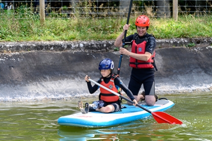 A boy with a prosthetic leg stands on a paddle board, on water. while an instructor kneels behind him