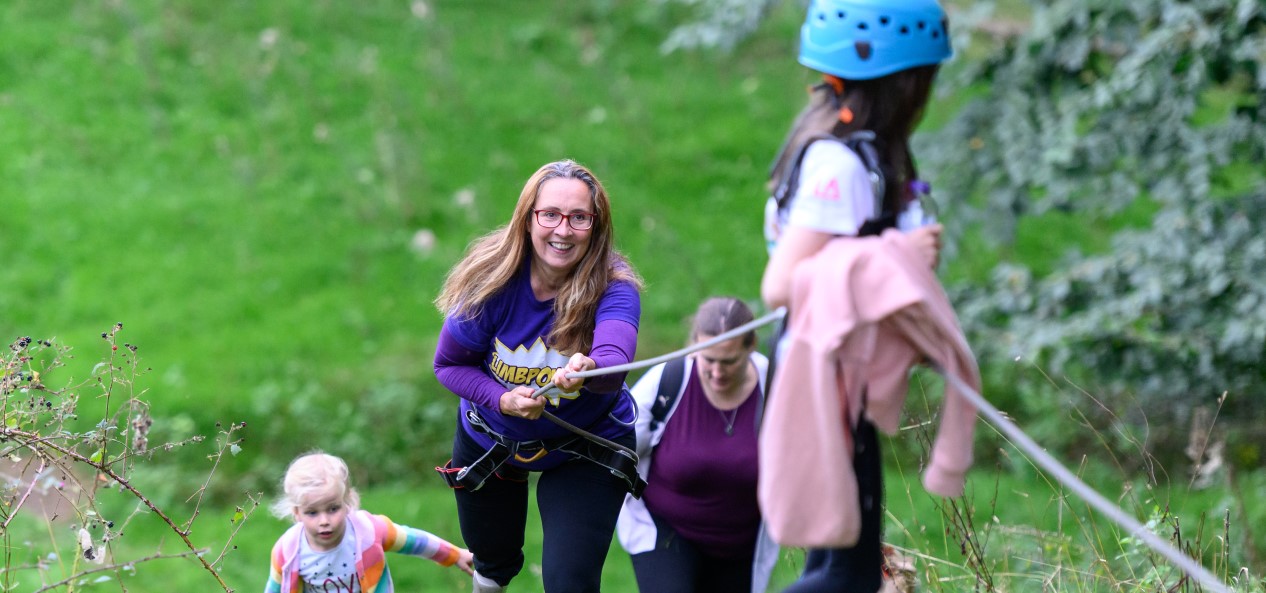 A woman smiles while pulling a rope to climb a hill. A girl in a helmet is at the top of the hill and there are other climbers below.