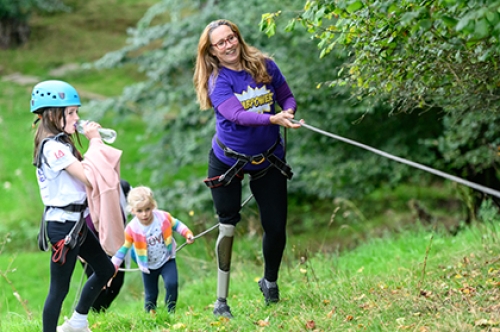 A woman with a prosthetic leg uses a rope to climb up a slope and two small girls watch on.