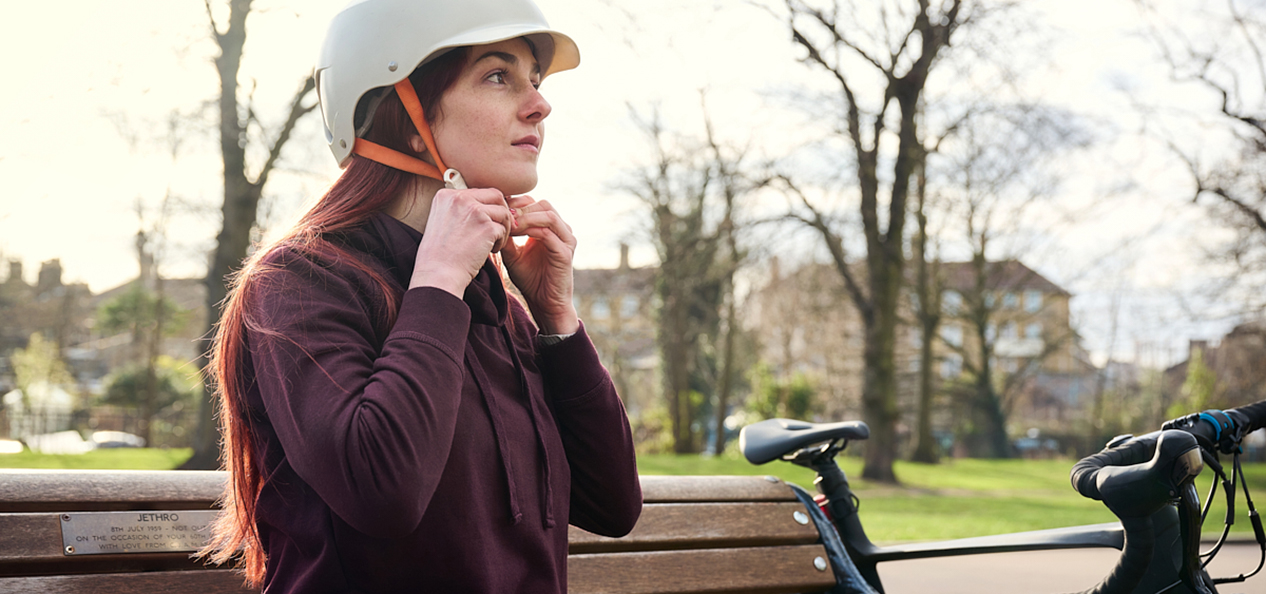 A woman, sitting on a bench in a park, fastens her cycling helmet as her bike leans up against the end of the bench.