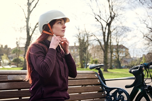 A woman, sitting on a bench in a park, fastens her cycling helmet as her bike leans up against the end of the bench.