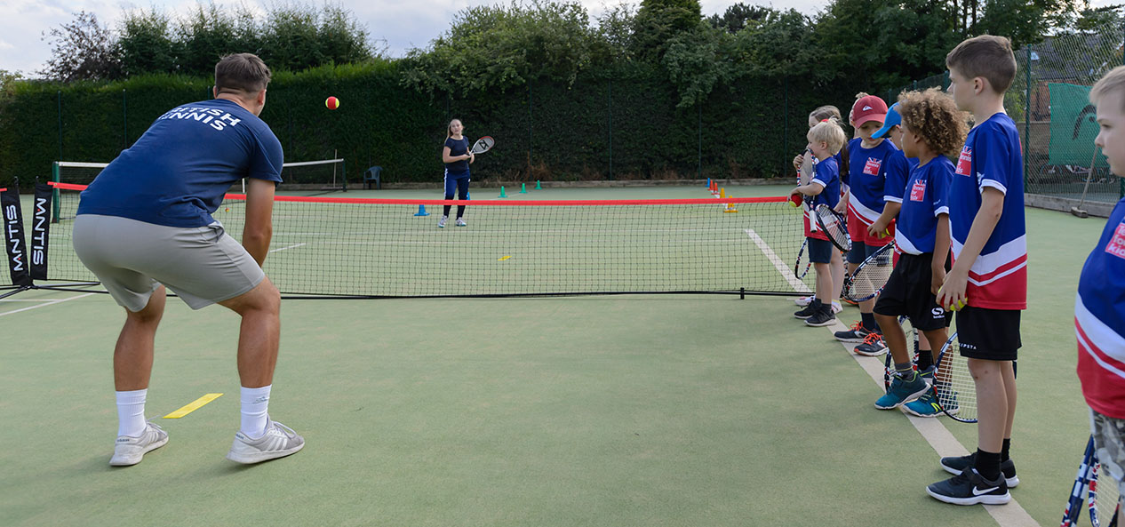 Children line up alongside a tennis court as a coach demonstrates a drill.