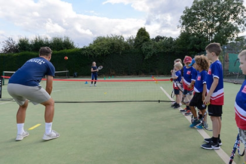 Children line up alongside a tennis court as a coach demonstrates a drill.