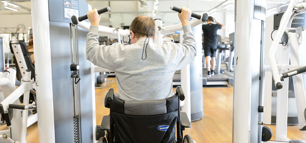 A man in a wheelchair uses a lat pull down machine in a gym.