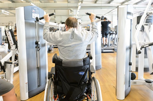 A man in a wheelchair uses a lat pull down machine in a gym.