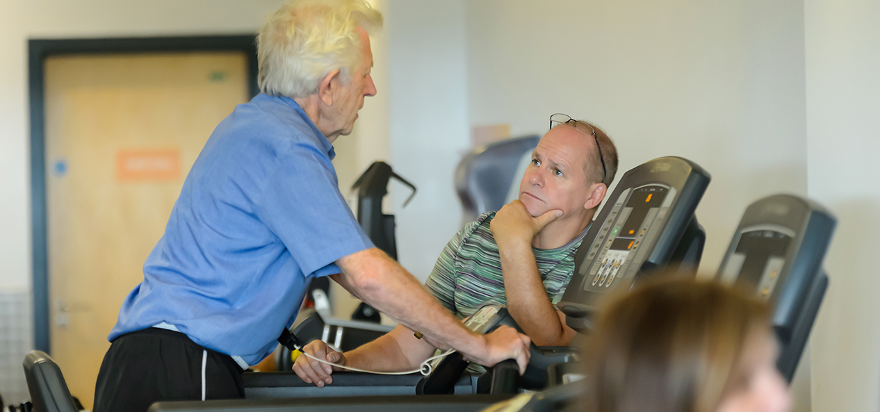 An old gentleman exercises on a walking machine indoors under the supervision of another man.