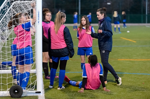 A female coach speaks to a teenage girls' football team at the side of an artificial football pitch