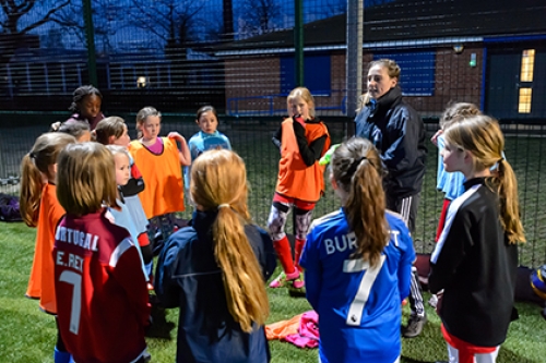 A coach gives instructions to a group of girls at a football coaching session on an artificial pitch