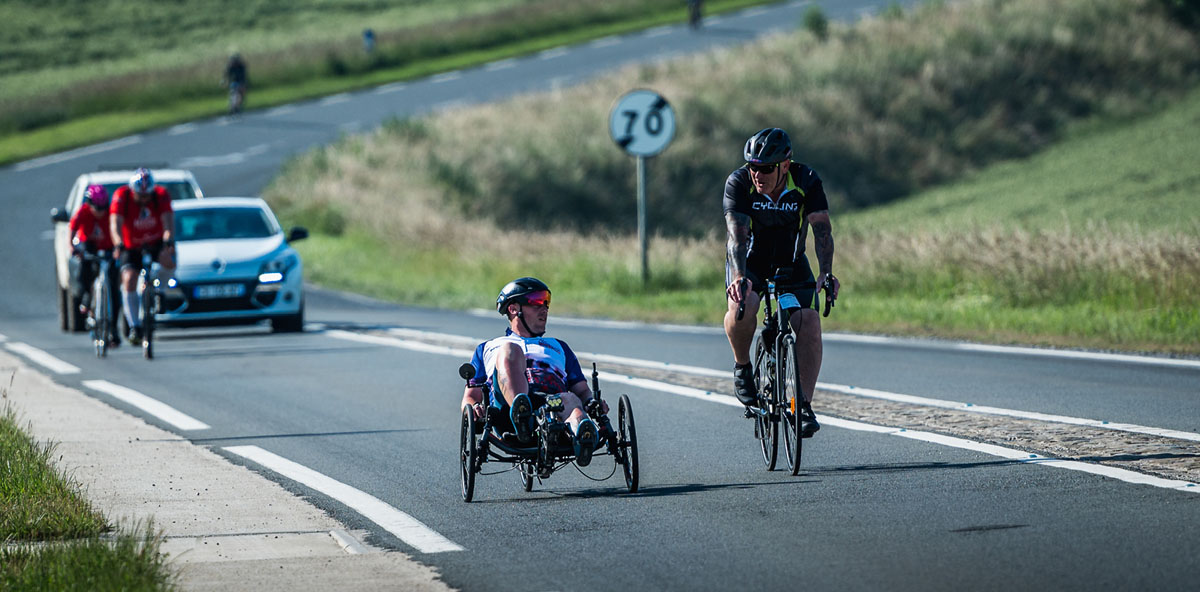 Two men are cycling up a hill and one of them is on an adapted bicycle.