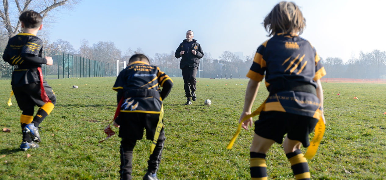 A coach instructs three young boys how to play tag rugby, on an open playing field