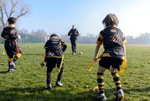 A coach instructs three young boys how to play tag rugby, on an open playing field