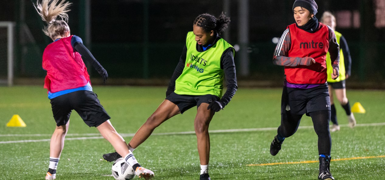 Two young women wearing football bibs compete for the ball on an outdoor artificial pitch at night, while a third watches on.