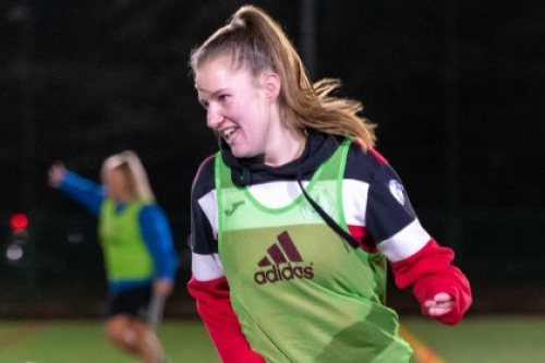 A young woman in a football bib smiles as she moves on an outdoor artificial pitch at night.