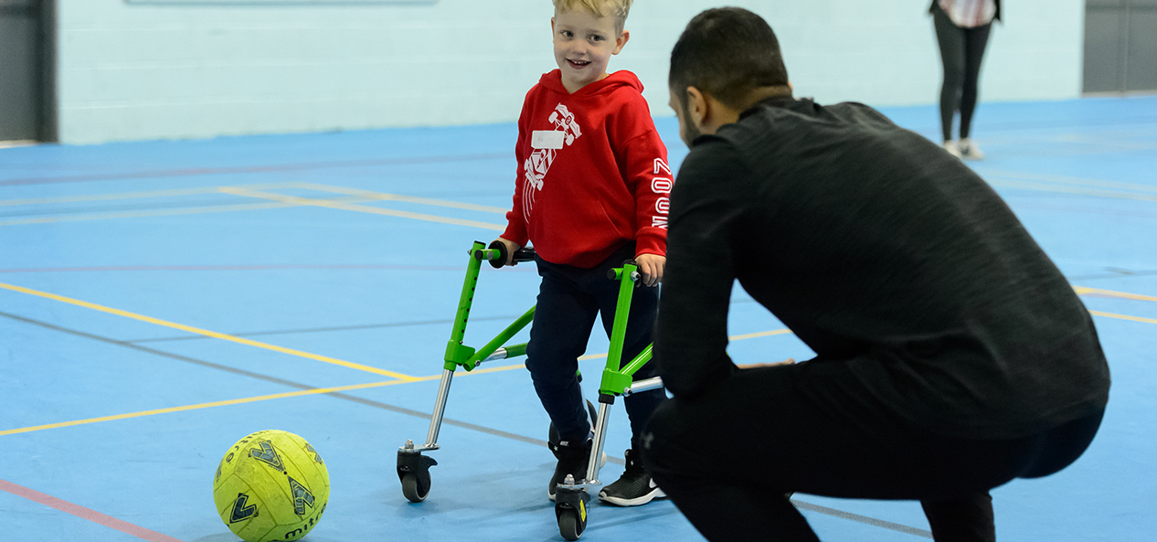 A boy using a walking frame plays football in a sports hall as a coach crouches down to talk to him