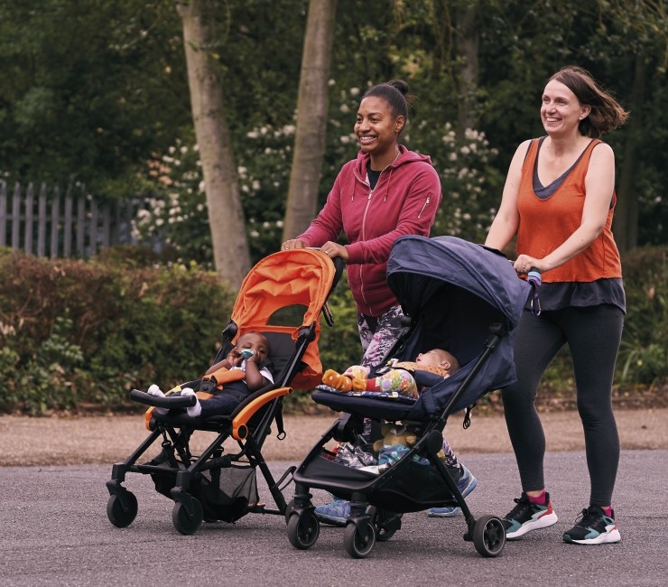 Two smiling women walk with their children in prams in a park.