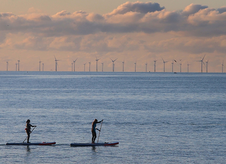 Two paddle boarders paddle in the sea, with a wind farm in the background