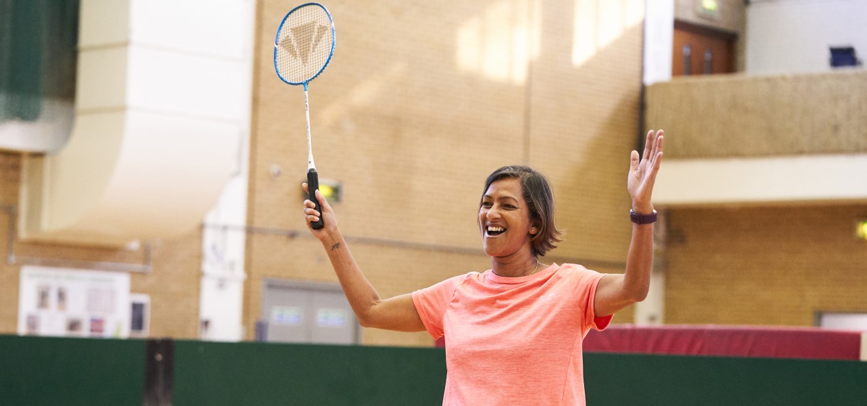 A smiling woman raises both arms, one of them holding a badminton racquet, on a court in a sports hall.