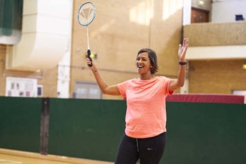 A smiling woman raises both arms, one of them holding a badminton racquet, on a court in a sports hall.