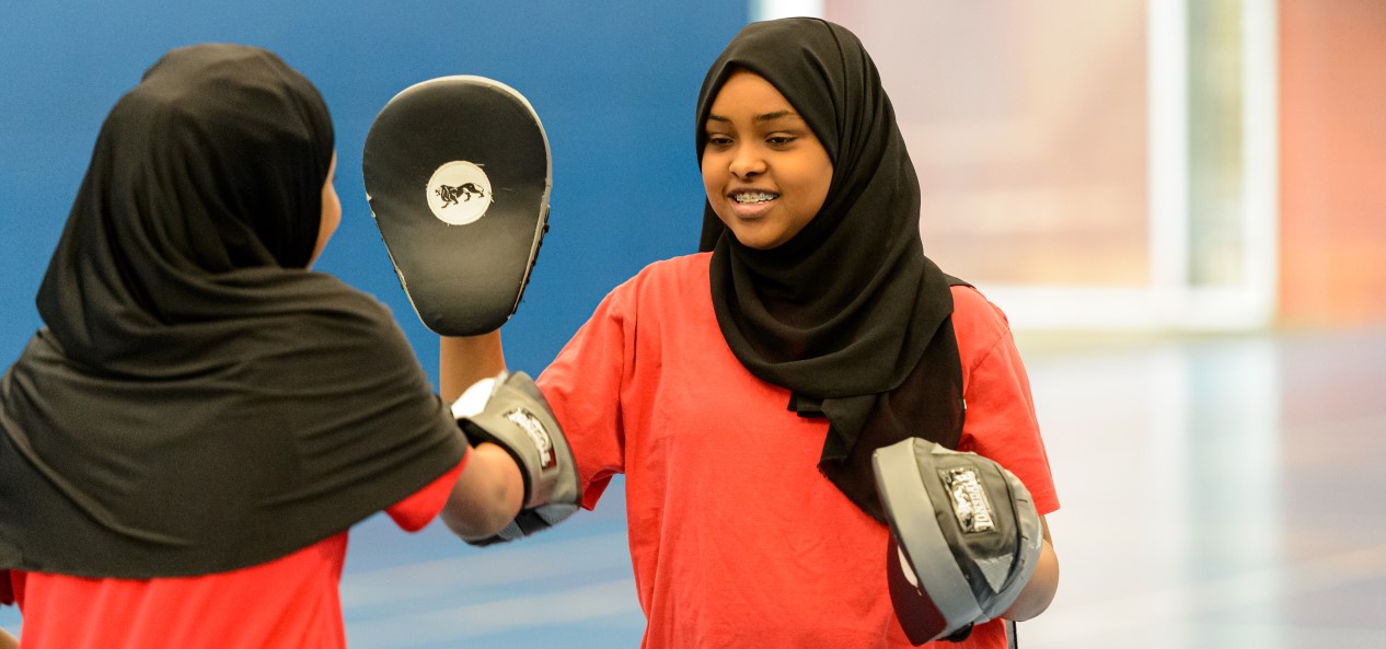 A young woman in a sports hall smiles while holding a boxing pad for another young woman to punch