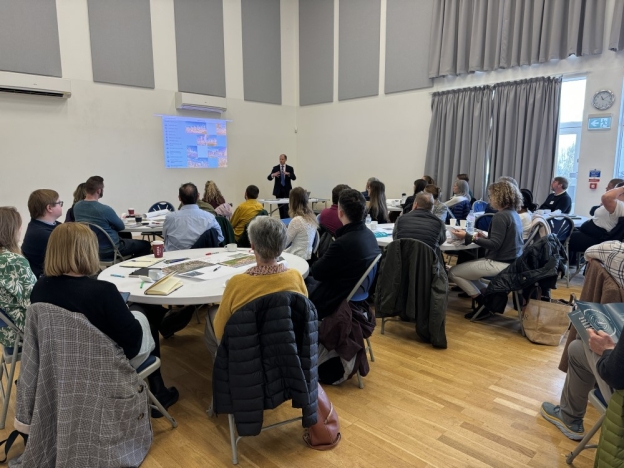 A man presents to people sat with documents at tables during a workshop.