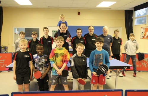 A group of young boys post in front of a table tennis table in a community centre, with their coaches standing behind the table.