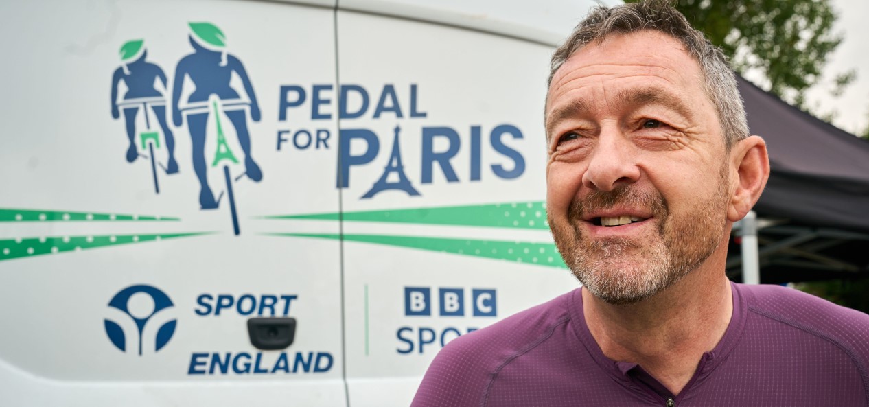 Chris Boardman wearing a Pedal for Paris cycling jersey in front of the Sport England electric vehicle for the campaign.