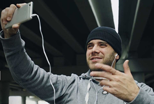 A man in a tracksuit top talks to his phone, while wearing headphones, delivering a sports session online.