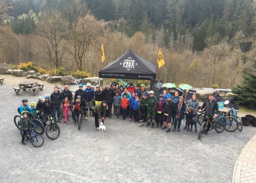 A large group of cyclists stand in front of a 'Trash Free Trails' gazebo in a forest