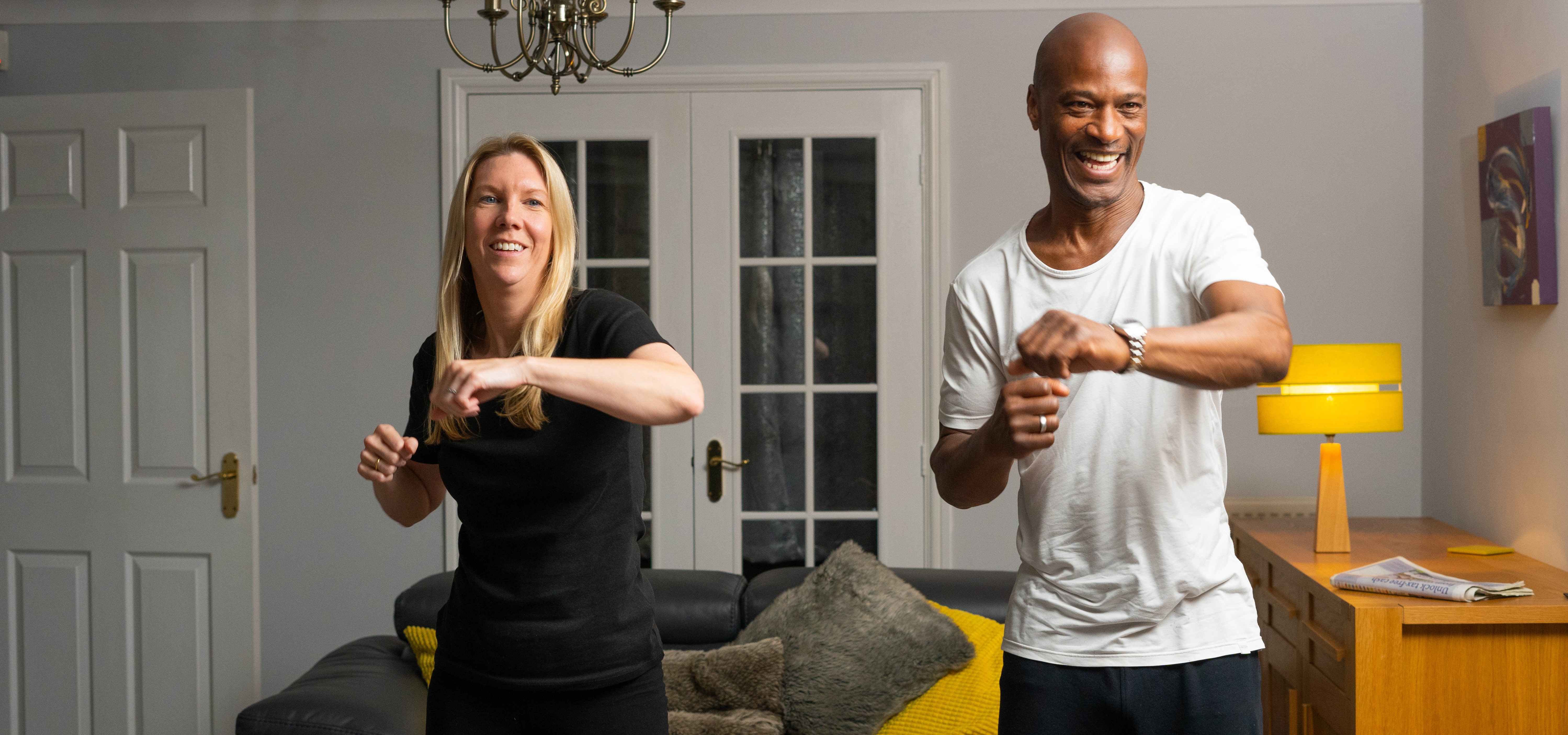 A smiling man and a woman stood side by side in a living room, taking part in a workout.