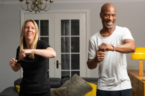 A smiling man and a woman stood side by side in a living room, taking part in a workout.